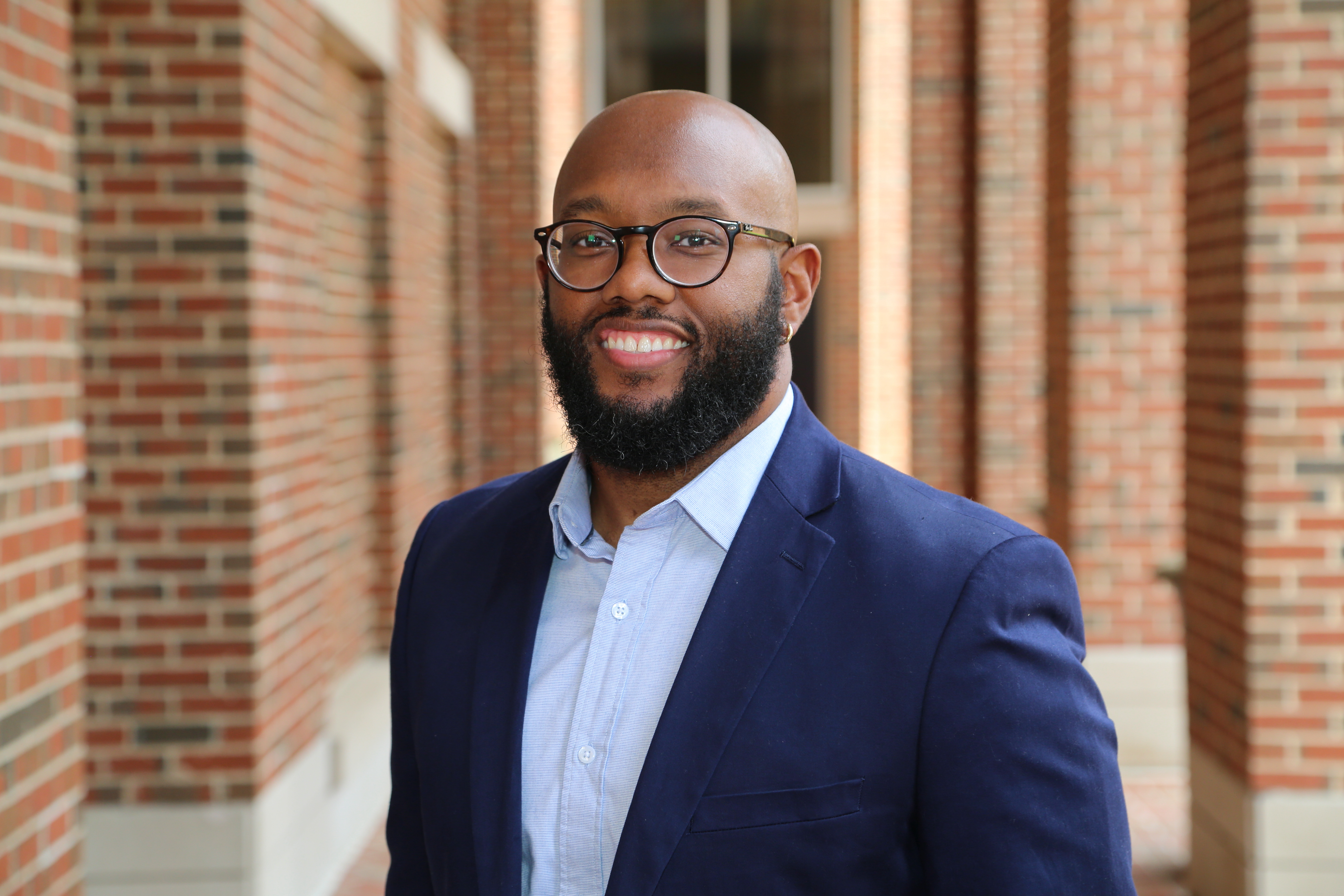 Dr. Morton wears glasses, a collared shirt, and a suit jacket while smiling outside a brick academic building.