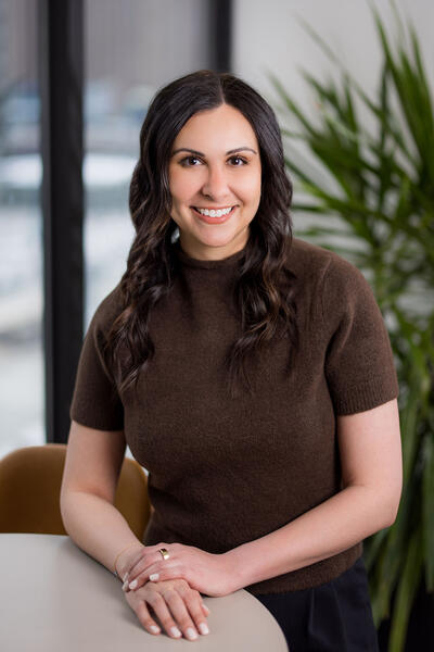 Sophia Odeh smiles and stands next to a chair with her hands placed in front of her on a table and a potted green palm behind her.