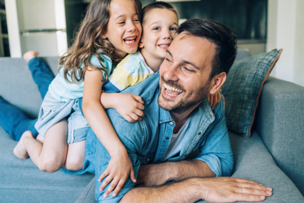 A father laughs and lies on a couch while two smiling children climb on his back