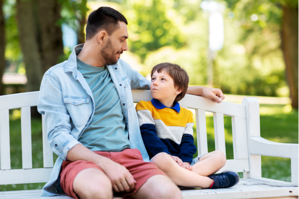 A smiling parent sits on a bench outdoors and looks down at their child sitting next to them. The child looks up at their parent while making a silly face.